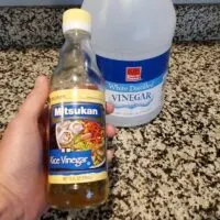 A Hand Holding Rice Vinegar With a Bottle of White Vinegar in the Background