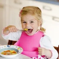 Blonde Toddler Eating a Spoonful of Vegetable Soup in a High Chair