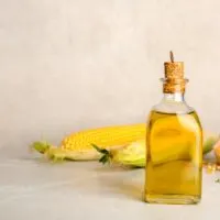 Bottle of corn oil and fresh cobs on table against light wall