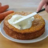 A woman's hand spreads frosting on a baked carrot cake with raw carrots in the background